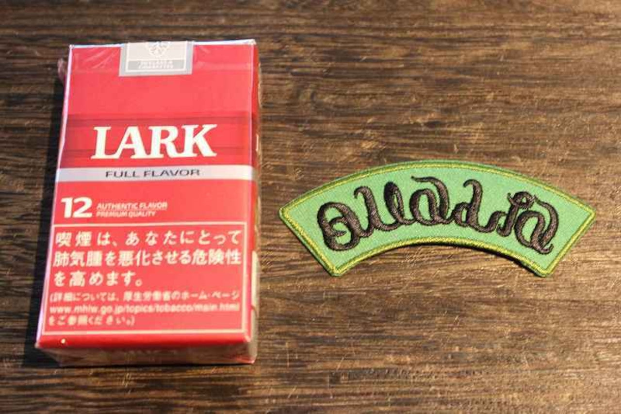 曲線的な黒い文字で刺繍されたアーチ型ワッペンの画像。緑色のベースに黄色い縁取りが施され、木製テーブル上に置かれている。ファッションやアウトドアグッズのカスタムに最適な刺繍ロゴワッペン