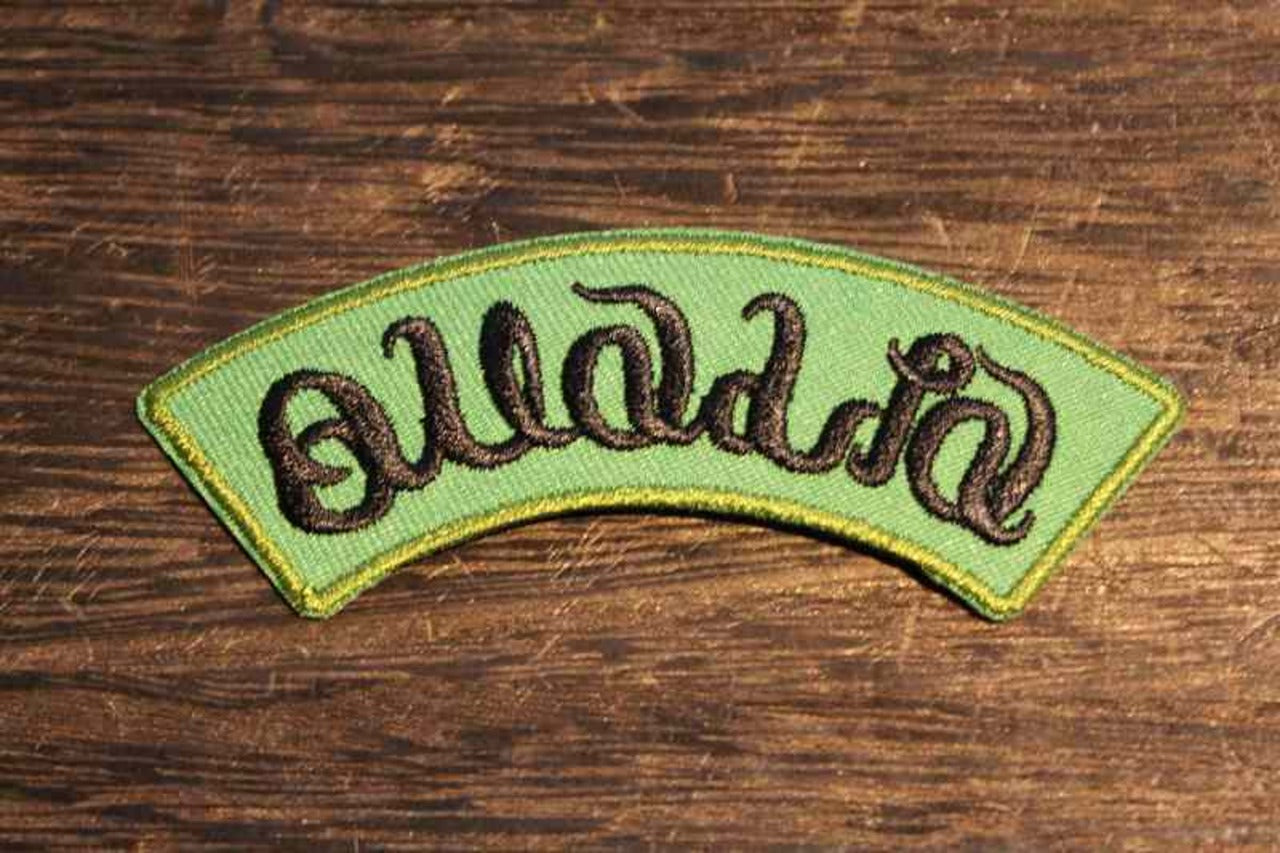 曲線的な黒い文字で刺繍されたアーチ型ワッペンの画像。緑色のベースに黄色い縁取りが施され、木製テーブル上に置かれている。ファッションやアウトドアグッズのカスタムに最適な刺繍ロゴワッペン2