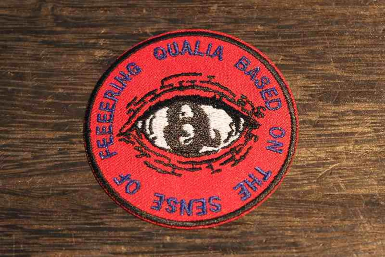 赤い背景に目のデザインと英語テキストが刺繍された丸型ワッペン。『QUALIA BASED ON THE SENSE OF FEELERING』の文字が円形に配置され、中央にはリアルな瞳の刺繍。個性的でインパクトのある刺繍ロゴパッチ