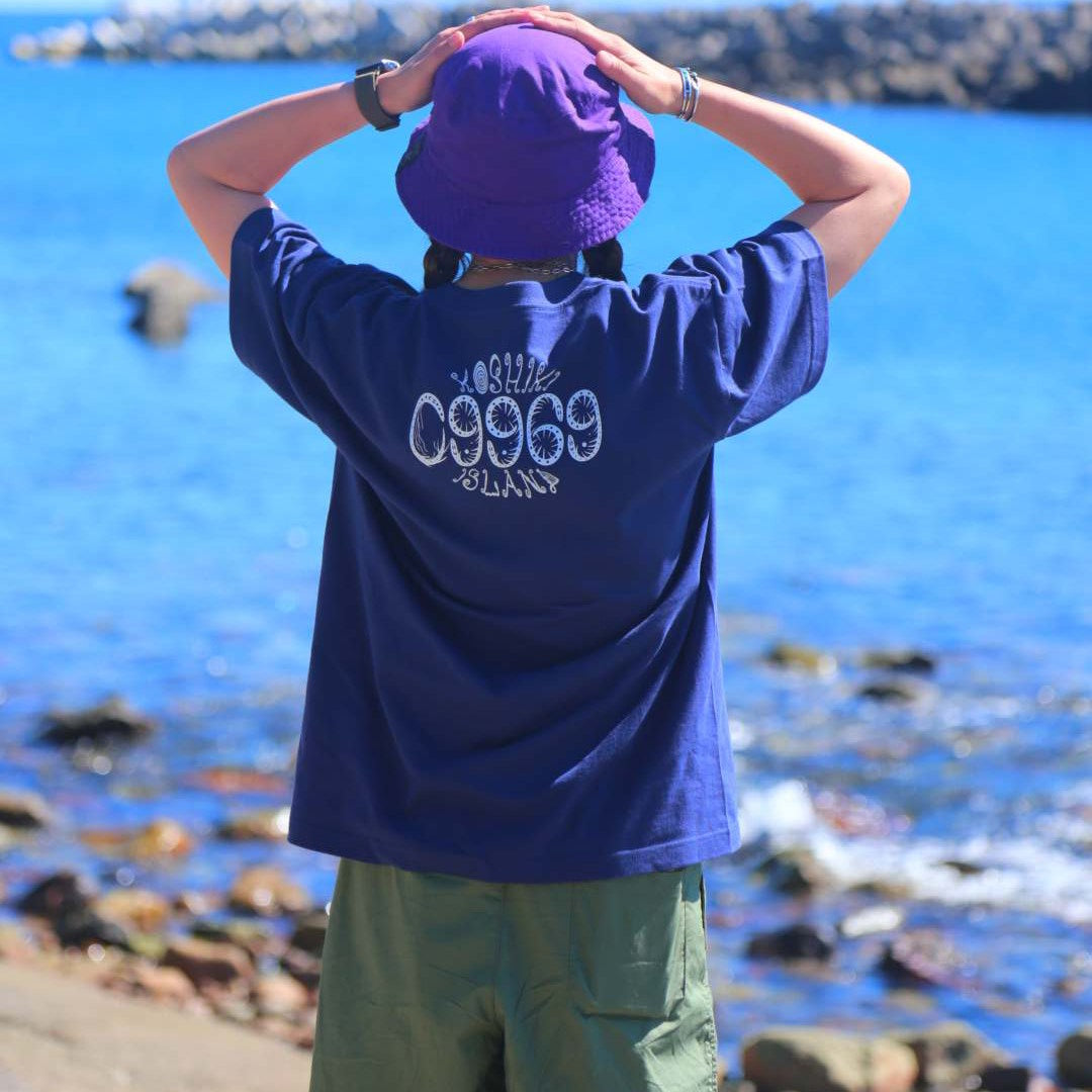 09969のロゴが背中に入ったネイビーのTシャツを着用。鮮やかな青い海を前に、背後から撮影された甑島の海辺に立つ女性