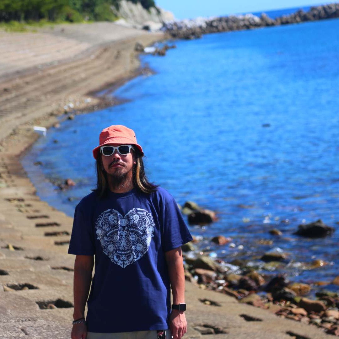 09969のネイビーTシャツに白いサングラスとオレンジのバケットハットを合わせた島スタイル。透き通る青い海と岩場を背景に、甑島の海辺に立つ男性の画像3