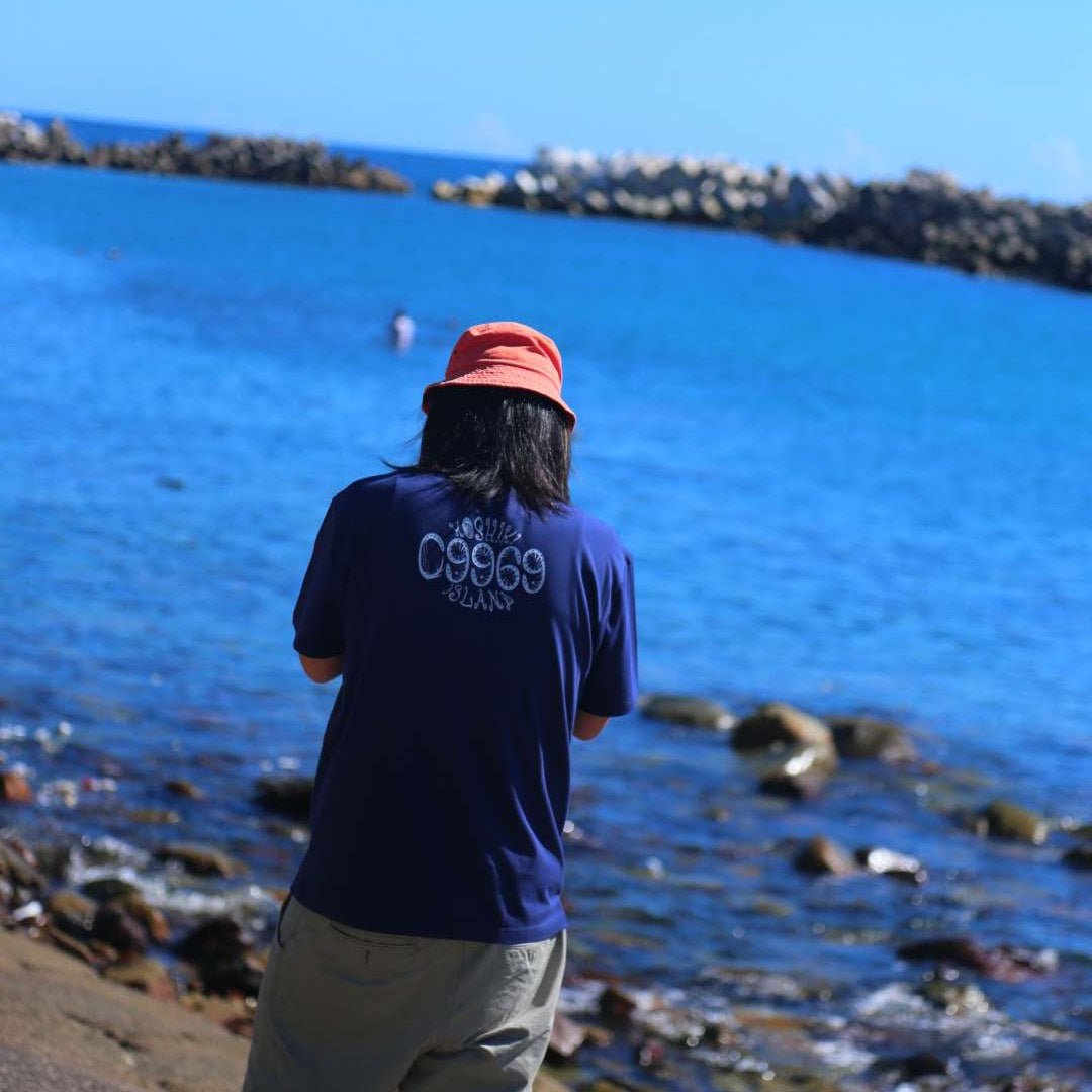 09969のロゴが背中に入ったネイビーのTシャツを着用。鮮やかな青い海を前に、背後から撮影された甑島の海辺に立つ男性