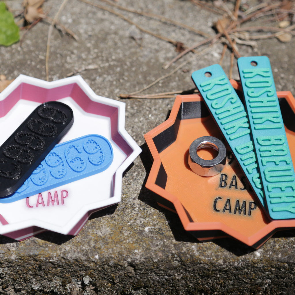 09969オリジナルのプレートやキーホルダーを、L.O.L BASE CAMPデザインの八角形トレイにまとめたカット。KOSHIKI BREWERYや09969ロゴ入りアイテムが並び、甑島発の観光・クラフトグッズとして展開