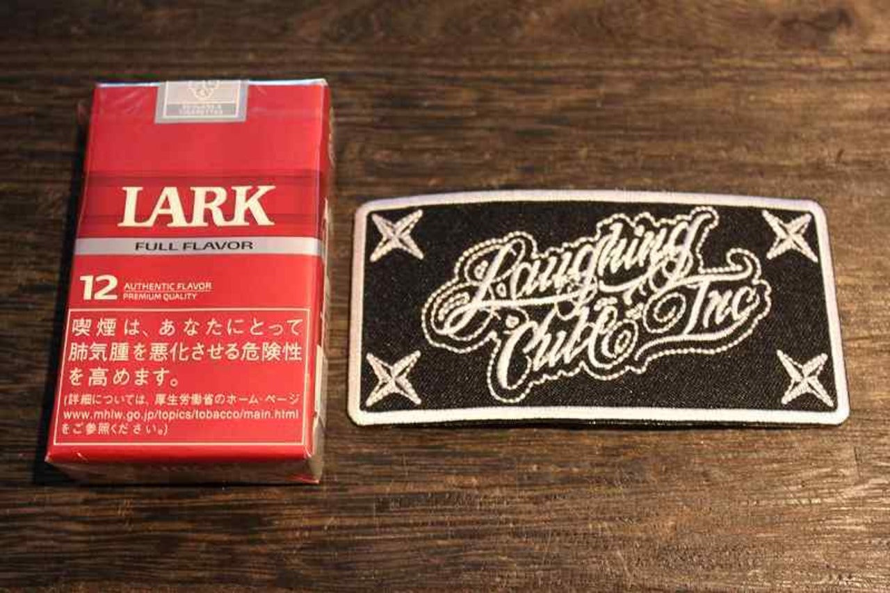 「Laughing Club Inc」の刺繍ワッペンの画像。黒地に白い刺繍でロゴ風の筆記体がデザインされ、四隅には手裏剣のような模様が配置されたクールなワッペンの画像1