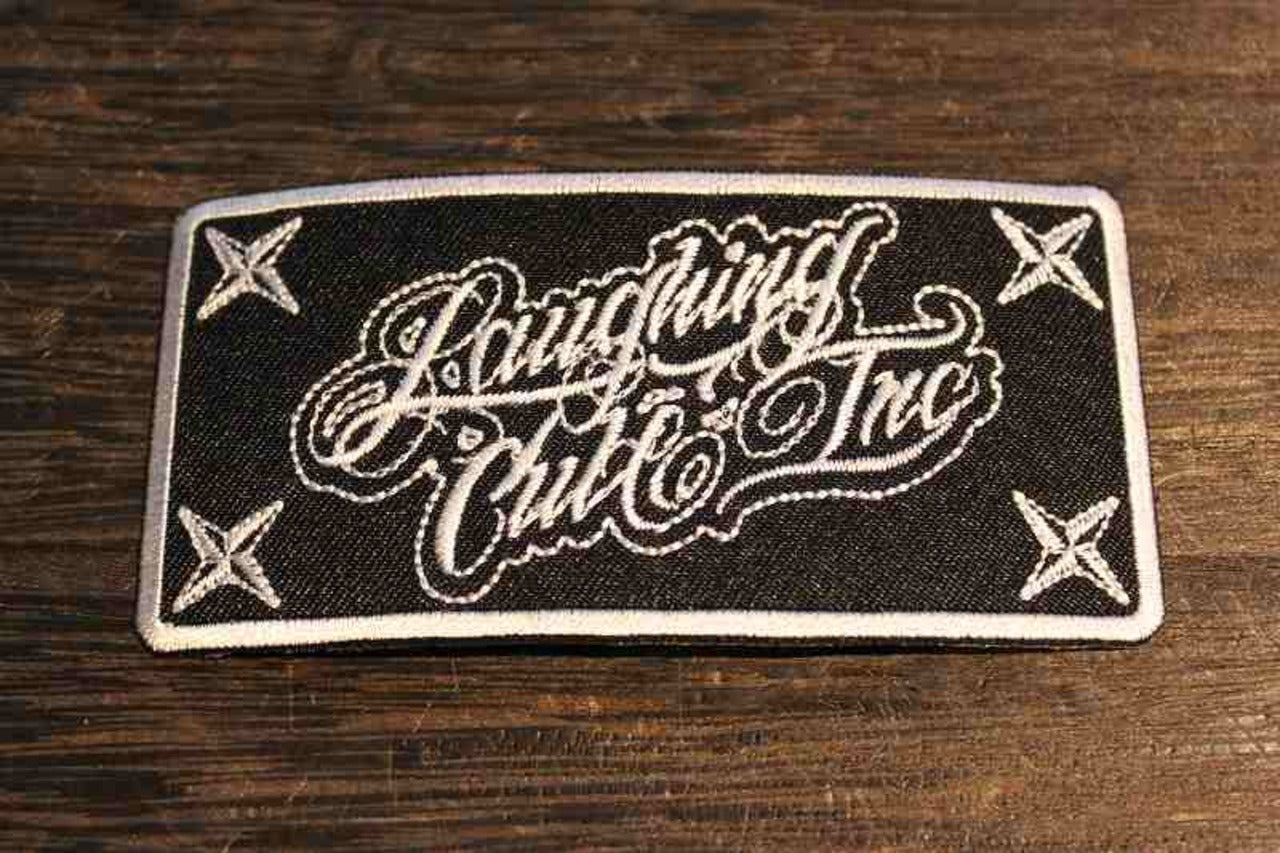 「Laughing Club Inc」の刺繍ワッペンの画像。黒地に白い刺繍でロゴ風の筆記体がデザインされ、四隅には手裏剣のような模様が配置されたクールなワッペンの画像2