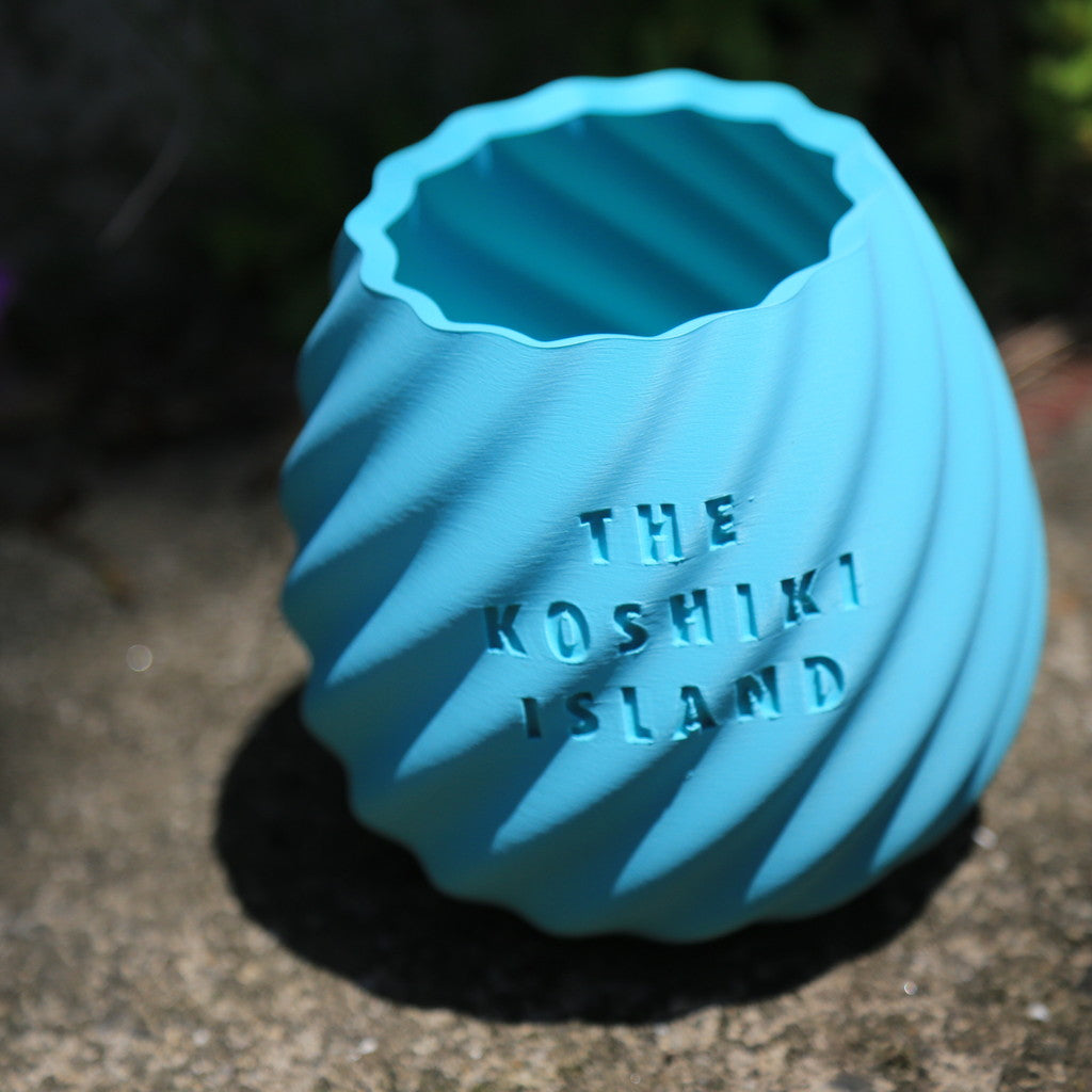 THE KOSHIKI ISLANDの文字が彫られた、水色のマットな質感の多角形カップ。ひねりのある立体形状が特徴で、09969。jpによる甑島オリジナルグッズの画像１